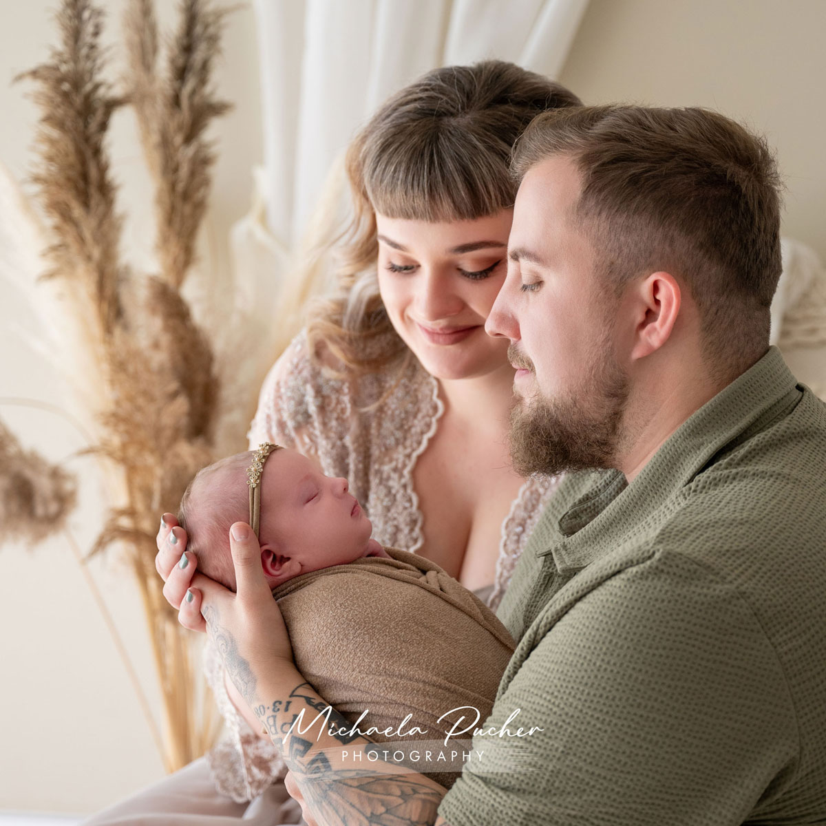 Junge Eltern halten ihr schlafendes Neugeborenes liebevoll im Arm; beide blicken zärtlich auf das Baby, umgeben von natürlichen Boho-Elementen.